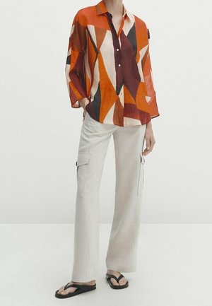 Person wearing a long-sleeve, multicolor geometric orange blouse, beige wide-leg cargo pants, and black flip-flops, standing against a white background.