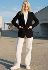 Smiling woman with long blonde hair stands outdoors wearing a black blazer, white shirt, wide white pants, and white sneakers under clear sky.