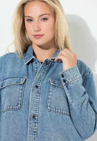 Lichtblauwe denim overhemd met drukknoopjes, twee borstzakken en contrasterende stiksels. De stof heeft een versleten textuur. Model past de kraag aan.