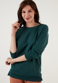 Felpa lunga teal con maniche a giro e polsini a costine. Caratterizzata da spacco laterale all'orlo per maggiore libertà di movimento. Realizzata in materiale morbido.