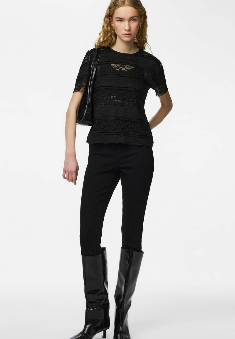 Jeune femme aux cheveux bouclés portant un haut en dentelle noire, un pantalon noir, des bottes noires montantes et un sac à bandoulière noir.