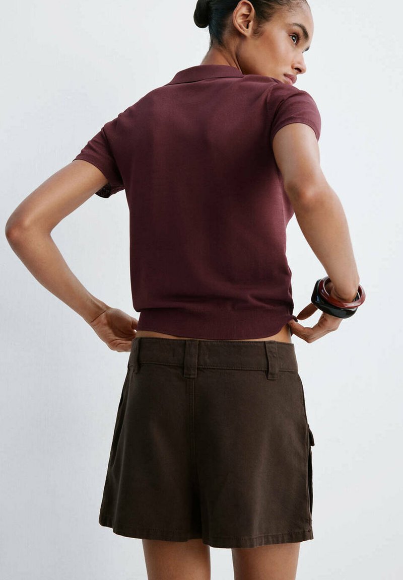 Femme aux cheveux en chignon, portant un haut bordeaux à manches courtes, un short marron foncé et plusieurs bracelets, ajustant son vêtement dans le dos.