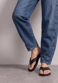Personne portant un jean bleu et des sandales noires, debout avec un pied légèrement levé sur un sol gris et un fond clair.