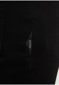 Un gros plan sur une poche latérale de pantalon en denim noir, avec une petite étiquette tissée affichant le nom de la marque "CHASIN". Texture lisse.