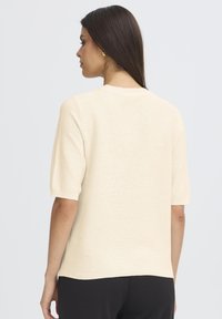 Heledbeez värvi lühikeste varrukatega kampsun, millel on ribatud tekstuur ja ümmargune kaelus, otsekohene alumine serv ja lahe lõige.