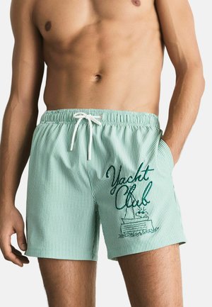 Gestreifte Badehose in Hellgrün mit elastischem Bund, weißem Kordelzug und "Yacht Club"-Grafik in dunklerem Grün.