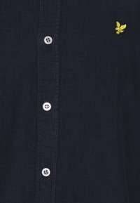 Ciemnoniebieska koszula sztruksowa z białymi guzikami i małym, żółtym haftowanym logo orła na piersi.