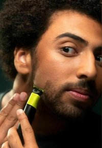 Black and green electric trimmer held near a person's cheek, featuring a yellow grip and a detailed shaving head for precision.