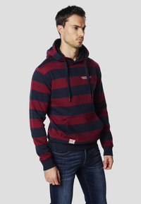 Mikina s kapucňou v bordovo-modrých pruhovaných farbách s predným vreckom a logom. Vyrobená z bavlny s rebrovanými manžetami a lemom. Nosí sa s tmavými džínsami.