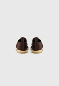 Pantofi din suede maro cu tăietură joasă, având o talpă din sfoară de iută. Designul include cusături duble și un mic sistem de prindere la spate pentru o îmbrăcare ușoară.