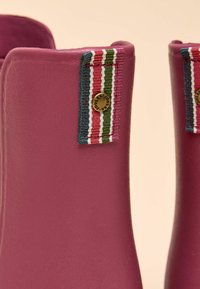 Fialové gumové holínky s texturovaným povrchem, zdobené vícenásobným pruhovaným látkovým akcentem a kulatým kovovým logem.