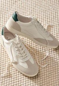 Paire de baskets blanches et beiges avec des languettes vertes au talon et des lacets défaits, reposant sur un tapis crème texturé.