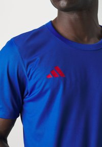 Blå atletisk t-shirt i mjukt tyg med en röd Adidas-logga på vänster bröst. Korta ärmar och rund halsringning.
