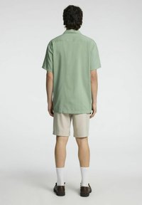 Camicia verde chiaro a maniche corte, vista posteriore, abbinata a pantaloni corti beige. La camicia presenta un colletto standard, vestibilità rilassata e una consistenza liscia.