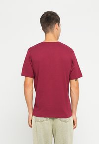 Burgundi pamut póló rövid ujjakkal, kerek nyakkal és egyszínű dizájnnal, világos színű, enyhén laza farmernadrággal viselve.