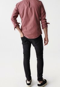 Camicia a maniche lunghe rossa a fantasia con polsini arrotolati, abbinata a jeans neri e sneaker neri. Cuciture visibili e dettagli di texture.