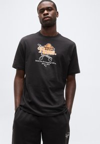 Νεαρός άνδρας που φοράει μαύρο μπλουζάκι Puma με γραφικά καλαθοσφαίρισης και καλαθιού αγορών και κείμενο, στέκεται με τα χέρια στις τσέπες.