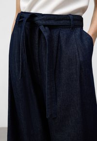 Mörkblå denimbyxor med vida ben, som har ett knytbälte i midjan och synliga sömnadsdetaljer längs kanterna.