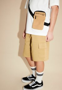 Bolso bandolera marrón con cierre de cremallera y correa negra, llevado sobre una camisa blanca y pantalones cortos beige. Zapatillas negras y calcetas a rayas completan el look.