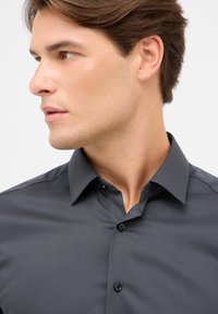 Camicetta grigia con bottoni, realizzata in tessuto texturizzato, colletto a punta e bottoni neri. La profilazione del modello mette in evidenza i dettagli del design della camicia.