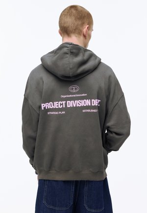 Moški s kratkimi lasmi, oblečen v temno sivo kapuco z roza napisom "Project Division" in temno modrimi kavbojkami, obrnjen stran, na belem ozadju.