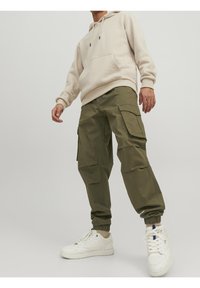 Man in een beige hoodie, olijfgroene cargobroek met elastische boorden, en witte sneakers die tegen een effen lichte achtergrond staat.