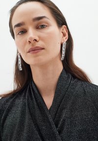 Top avvolgente nero e scintillante con scollatura a V, realizzato in tessuto texture e accompagnato da grandi orecchini metallici rettangolari con accenti di cristallo.