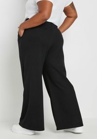 Svarta vida byxor med hög midja, med sidofickor och en slät textur. Kombinerade med vita skor som visar en ankel detalj.