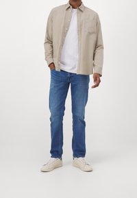 Béžová košile na knoflíky přes bílé tričko, v kombinaci s modrými džínami a světle zbarvenými teniskami. Ležérní střih s detailem náprsní kapsy.