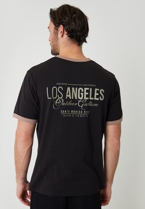 Homme de dos portant un t-shirt noir avec une bordure beige, texte « Los Angeles Outdoor Culture Santa Monica Ride » imprimé au dos.