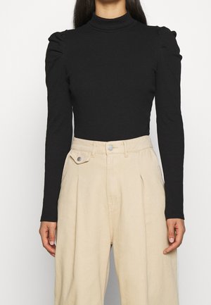 Femme portant un haut noir cintré à manches longues avec des épaules bouffantes et un pantalon beige ample taille haute.