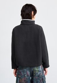 Schwarzer Fleece-Pullover mit hohem Kragen, lockerer Passform und strukturiertem Material. Verfügt über ein gesticktes Patch im oberen Rückenbereich und sitzende Schultern.