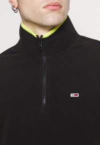 Svart fleece halvdraget pullover med en neongul krage och en liten broderad logotyp. Har en främre dragkedja och strukturerad tyg.