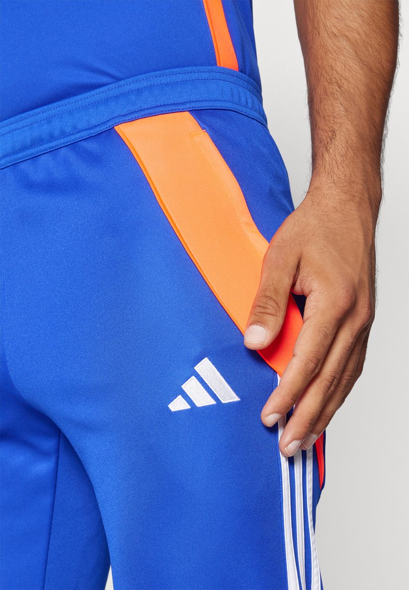 Blaue Sporthose mit orangefarbenen Seitenelementen, ausgestattet mit dem weißen adidas-Logo und drei weißen Streifen entlang der äußeren Naht. Glatte Textur.