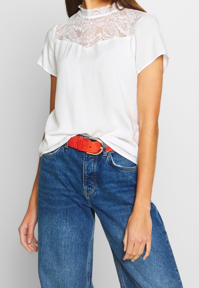 Femme portant un haut à manches courtes avec empiècement en dentelle blanche, rentré dans un jean bleu avec une ceinture orange vif, debout devant un fond clair uni.