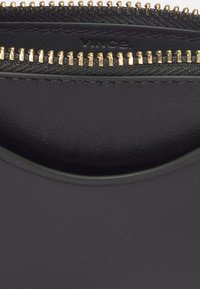 Gros plan sur une surface en cuir noir avec deux fermetures éclair en métal doré et une partie du nom de la marque embossé « Vince ».