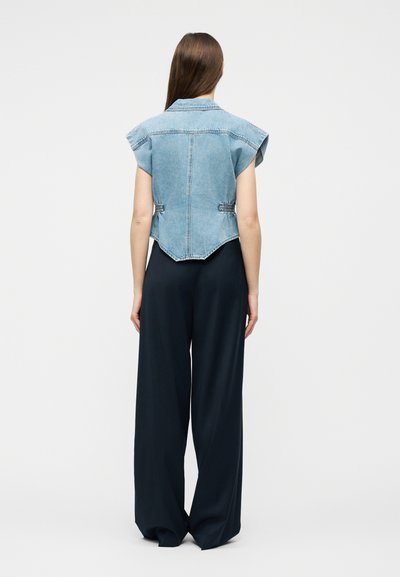 Femme aux longs cheveux bruns portant un gilet en denim sans manches et un pantalon large noir, debout de dos sur un fond uni.