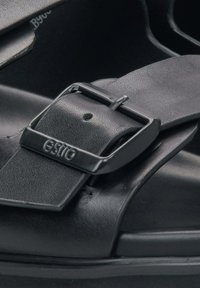 Černé sandály s texturovaným popruhem, čtvercovou černou přezkou a hladkou podrážkou. Kovové prvky mají minimalistický design a jsou vyrobeny z odolného materiálu.