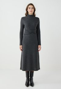 Graues Strickkleid mit hohem Kragen, langen Ärmeln, einem gedrehten Gürteldetail in der Taille und einem fließenden, knielangen Rock. Schwarze Ankle-Boots.