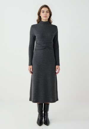 Graues Strickkleid mit hohem Kragen, langen Ärmeln, einem gedrehten Gürteldetail in der Taille und einem fließenden, knielangen Rock. Schwarze Ankle-Boots.