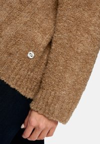 Brauner strukturierten Strickpullover mit einem gerippten Saum und einem metallischen Logo-Knopfdetail auf der linken Seite. Der Pullover hat eine weiche, flauschige Oberfläche.