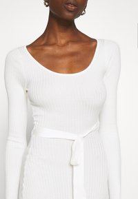 Abito lungo a maniche lunghe bianco con scollatura a cuore e cintura in tessuto annodata in vita, caratterizzato da una texture liscia e un design aderente.