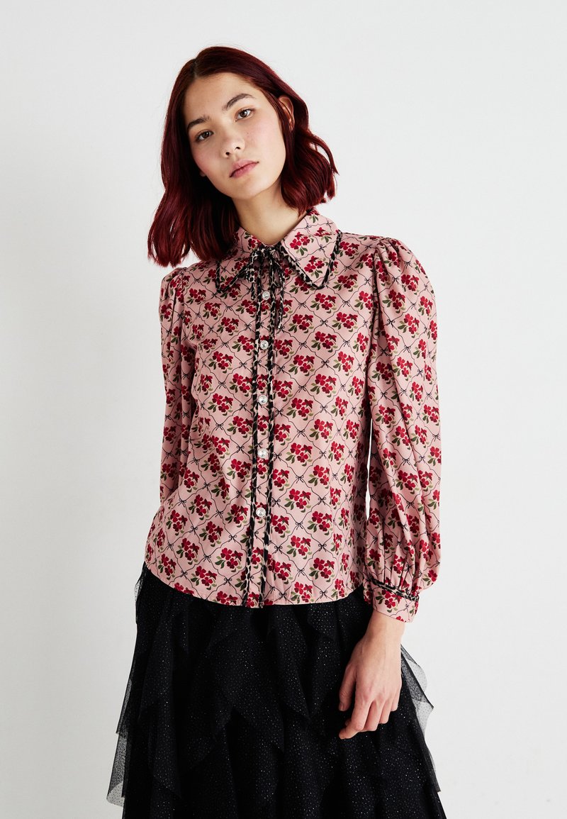 Blusa rosa con motivo floreale, maniche a sbuffo, chiusura con bottoni e rifiniture nere. Abbinata a una gonna in tulle nero a strati.