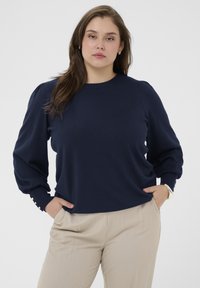 Marineblauwe pullover trui met lange, pofmouwen en knoopaccenten bij de polsen, gecombineerd met licht beige broeken.