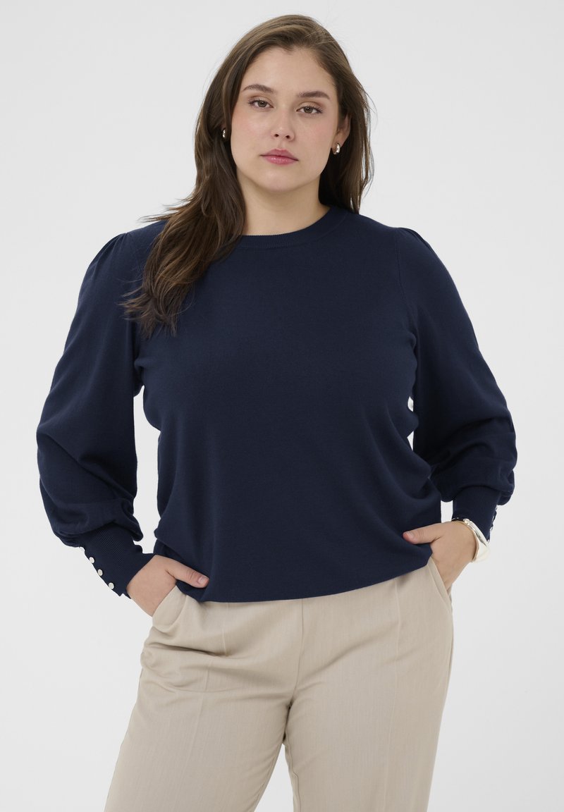 Marineblauwe pullover trui met lange, pofmouwen en knoopaccenten bij de polsen, gecombineerd met licht beige broeken.