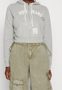 Felpa grigia con zip e stampa bianca "BDG JEANS" e "98", dotata di cappuccio regolabile e tasche a marsupio a contrasto, abbinata a pantaloni cargo color verde oliva.