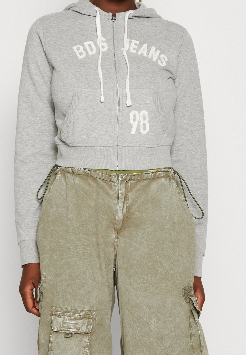 Felpa grigia con zip e stampa bianca "BDG JEANS" e "98", dotata di cappuccio regolabile e tasche a marsupio a contrasto, abbinata a pantaloni cargo color verde oliva.