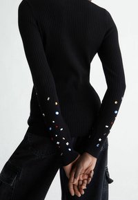 Pull noir côtelé à manches longues avec des ornements géométriques colorés aux poignets. Ourlet non fini et coupe ajustée.