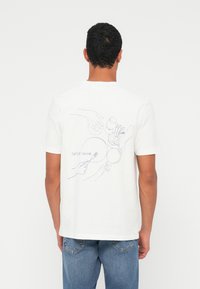 Man draagt een wit T-shirt met een lijntekening van handen die borden, een glas en een vork vasthouden, met de tekst "SAMSØE SAMSØE" op de achterkant.