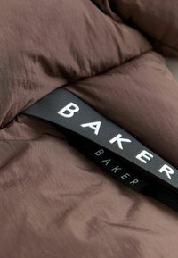 Ruda pūkinė striukė su lygiu paviršiumi ir šiek tiek blizgesio. Juoda etiketė su balta "BAKER" prekių ženklu aiškiai matoma.